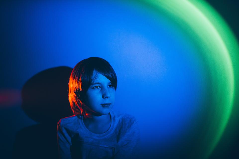 Child looks thoughtful, gazing into distance, with colorful light casting blue and red hues on the wall behind