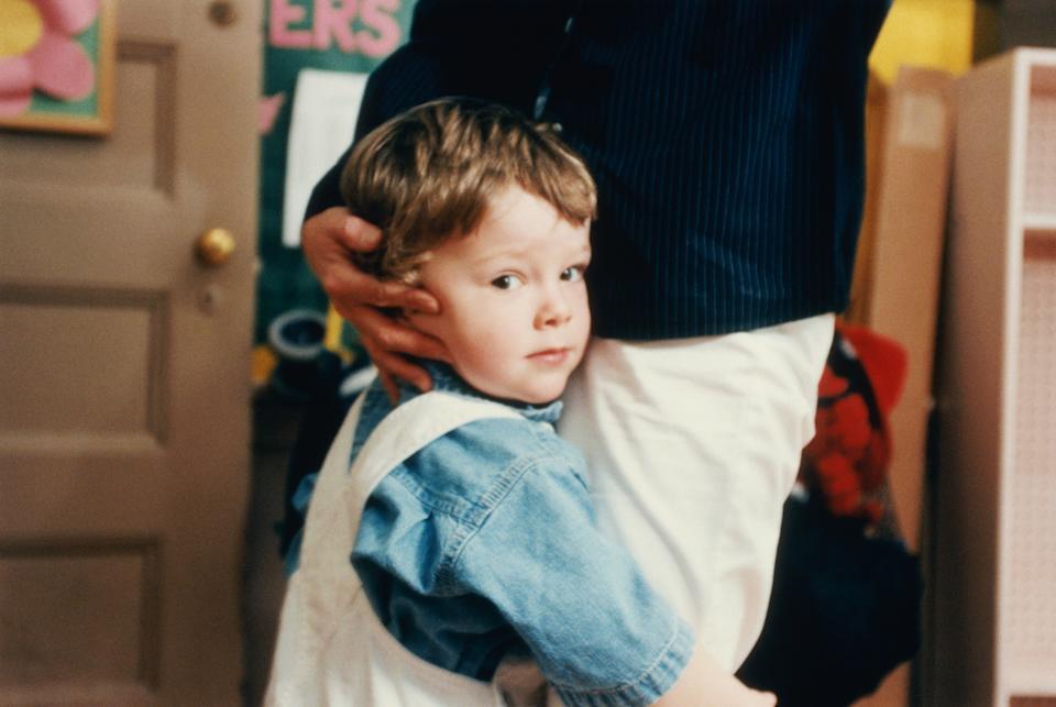 Child holding an adult's leg, looking to the side. The adult is partially visible, wearing a dark shirt and light pants. Classroom setting in the background