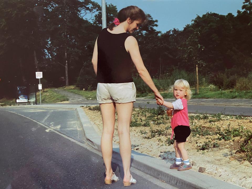 A woman in a sleeveless top and shorts holds hands with a young child with short hair on a quiet street, surrounded by greenery