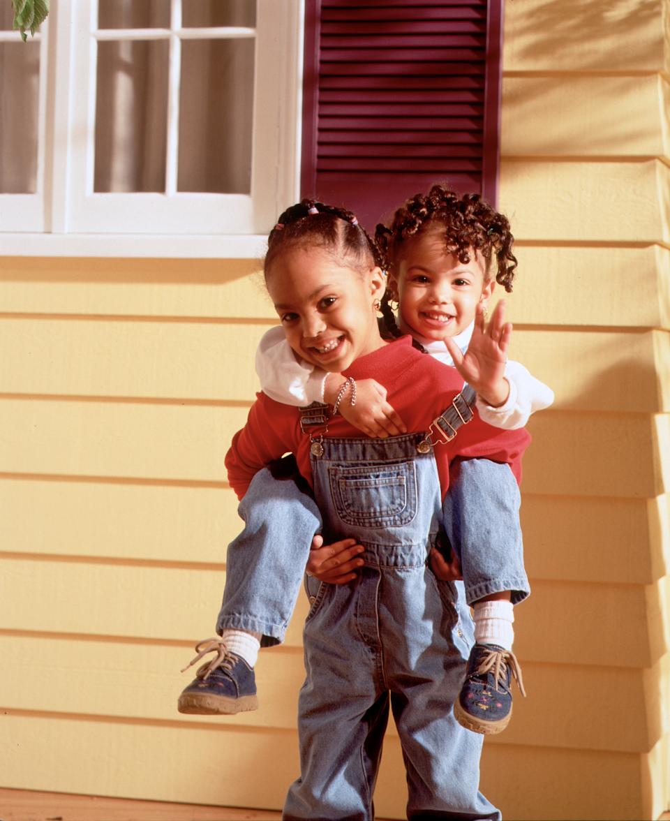 Two children smiling. One gives a piggyback ride to the other outside a house. Both wear casual outfits with denim overalls and shirts