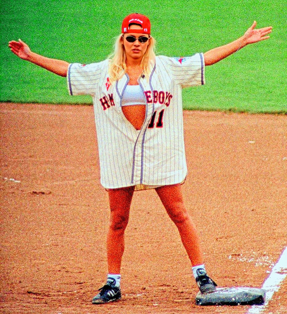 Person on a baseball field wearing a loose-fitting jersey, shorts, and cap, gesturing with arms wide open