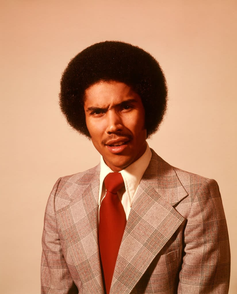 Person with an afro and mustache, wearing a plaid suit with wide lapels and a solid tie, looking directly at the camera