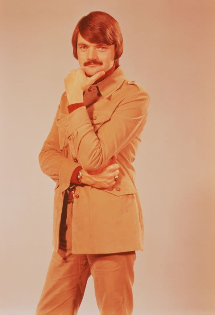 Person posing with hand on chin, wearing a safari-style jacket, long sleeves, and pants, with a thoughtful expression