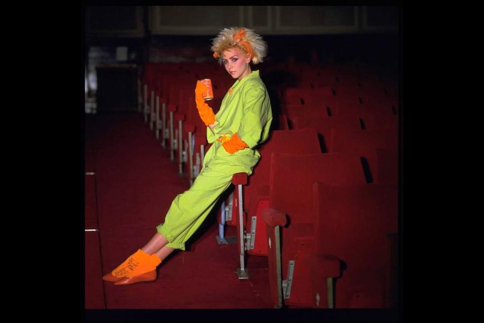 Person in avant-garde outfit sitting sideways on theater seat, wearing vibrant, eclectic clothing and striking a pose