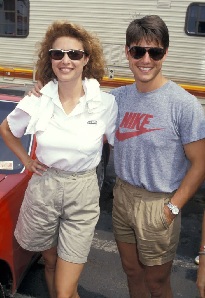 Two individuals stand smiling outdoors, both in casual attire with sunglasses. One wears a branded shirt, the other has a towel over their shoulder